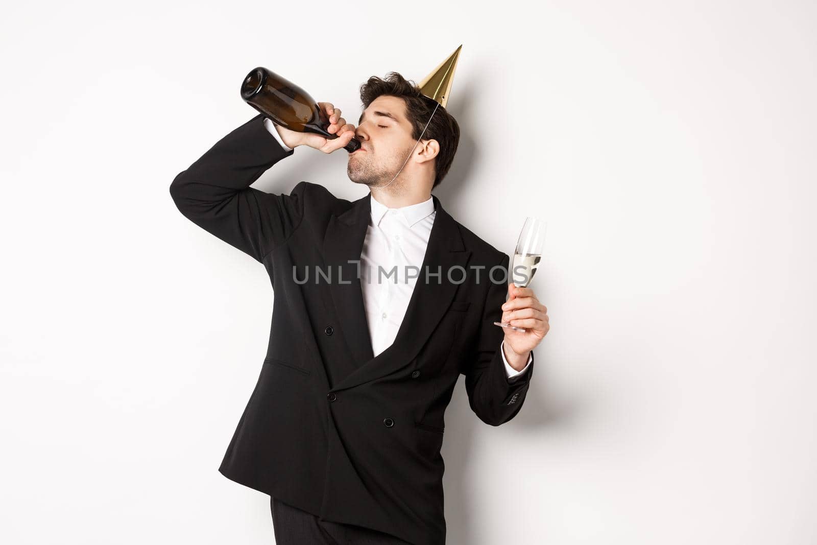 Image of handsome guy in trendy suit, drinking champagne from bottle and celebrating holiday, having a birthday party, getting drunk while standing over white background.