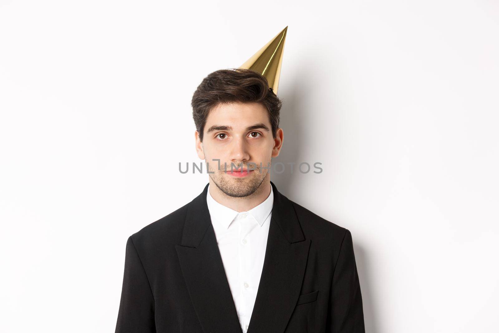 Close-up of handsome man in party hat and trendy suit, celebrating new year, standing against white background.