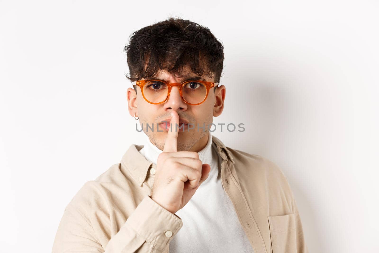 Close-up of grumpy young guy in glasses frowning and shushing, make taboo gesture with finger shuting lips, tell to be quiet, standing angry on white background by Benzoix