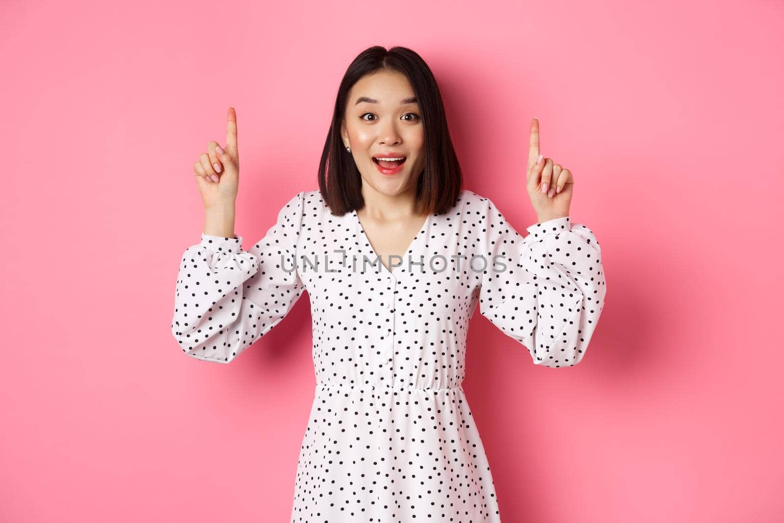 Amazed brunette asian girl pointing fingers up, showing advertisement, demonstrate spring promo offer, standing over pink background.