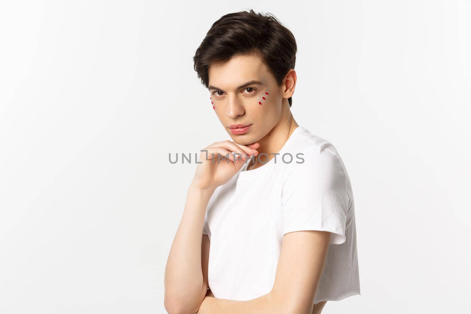 People, lgbtq and beauty concept. Beautiful androgynous male model with glitter on face, wearing crop top, looking with piercing eyes at camera, standing over white background.