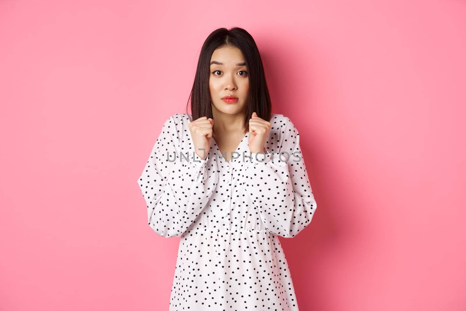 Scared cute asian girl in dress looking worried, feeling frightened and staring at camera nervously, standing over pink background.