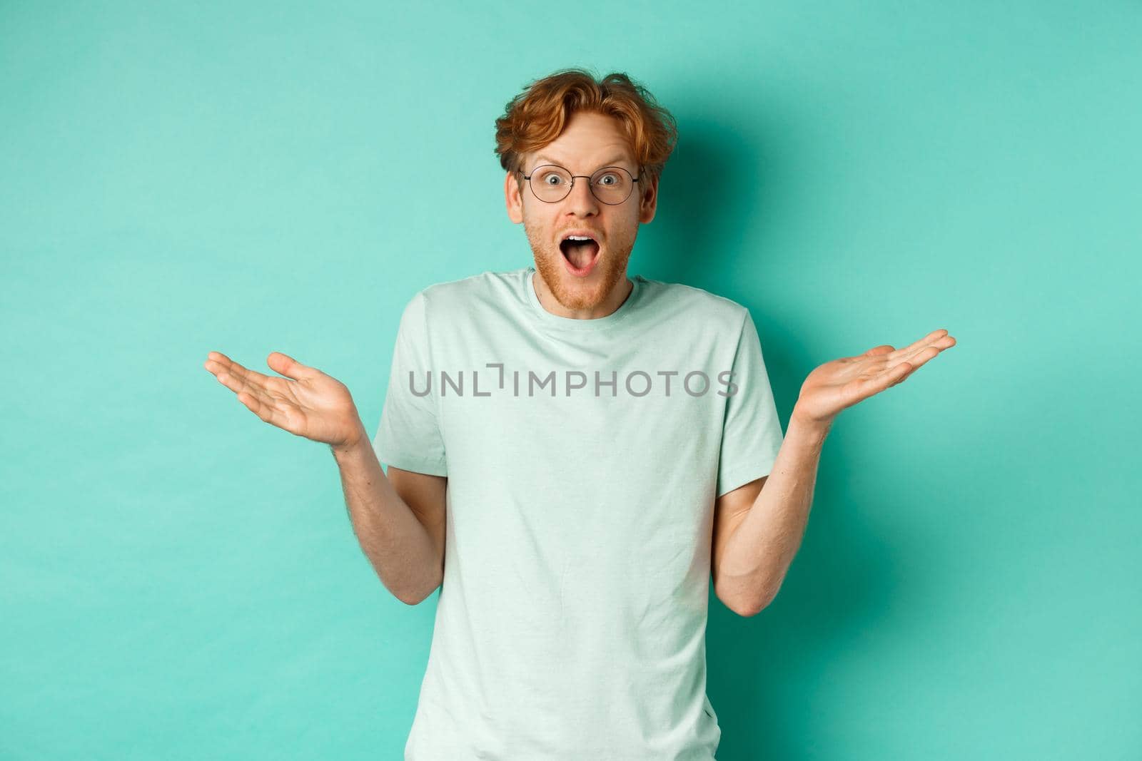 Surprised handsome man with red hair, wearing glasses, spread hands sideways and gasping in awe, looking wondered at camera, standing over turquoise background.