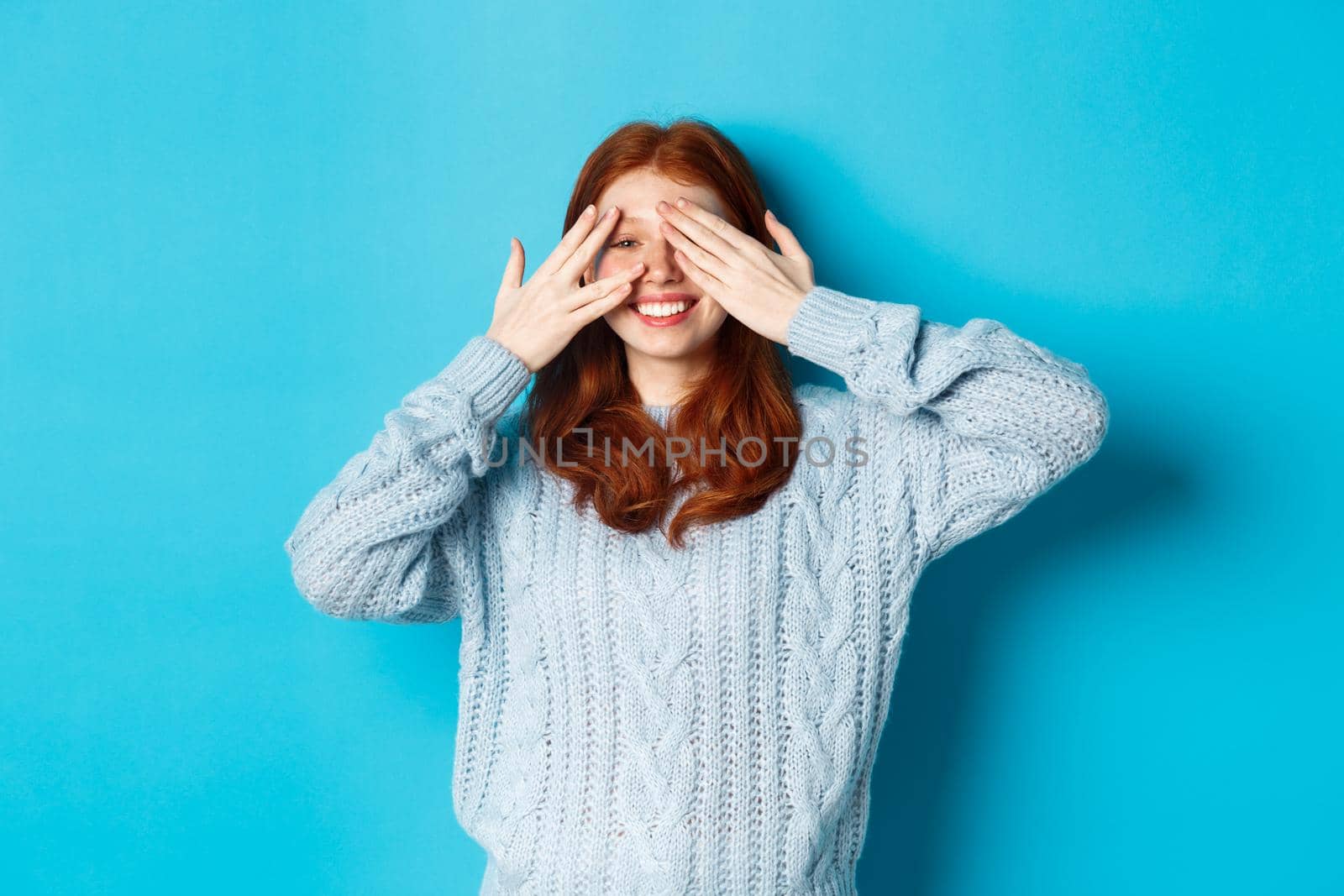 Excited teenage redhead girl open eyes to see holiday surprise, receiving presents, looking amazed at camera, standing over blue background.