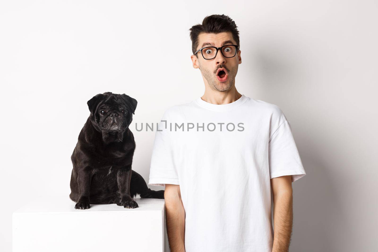 Image of dog owner and cute black pug looking at camera surprised and amazed, standing over white background.