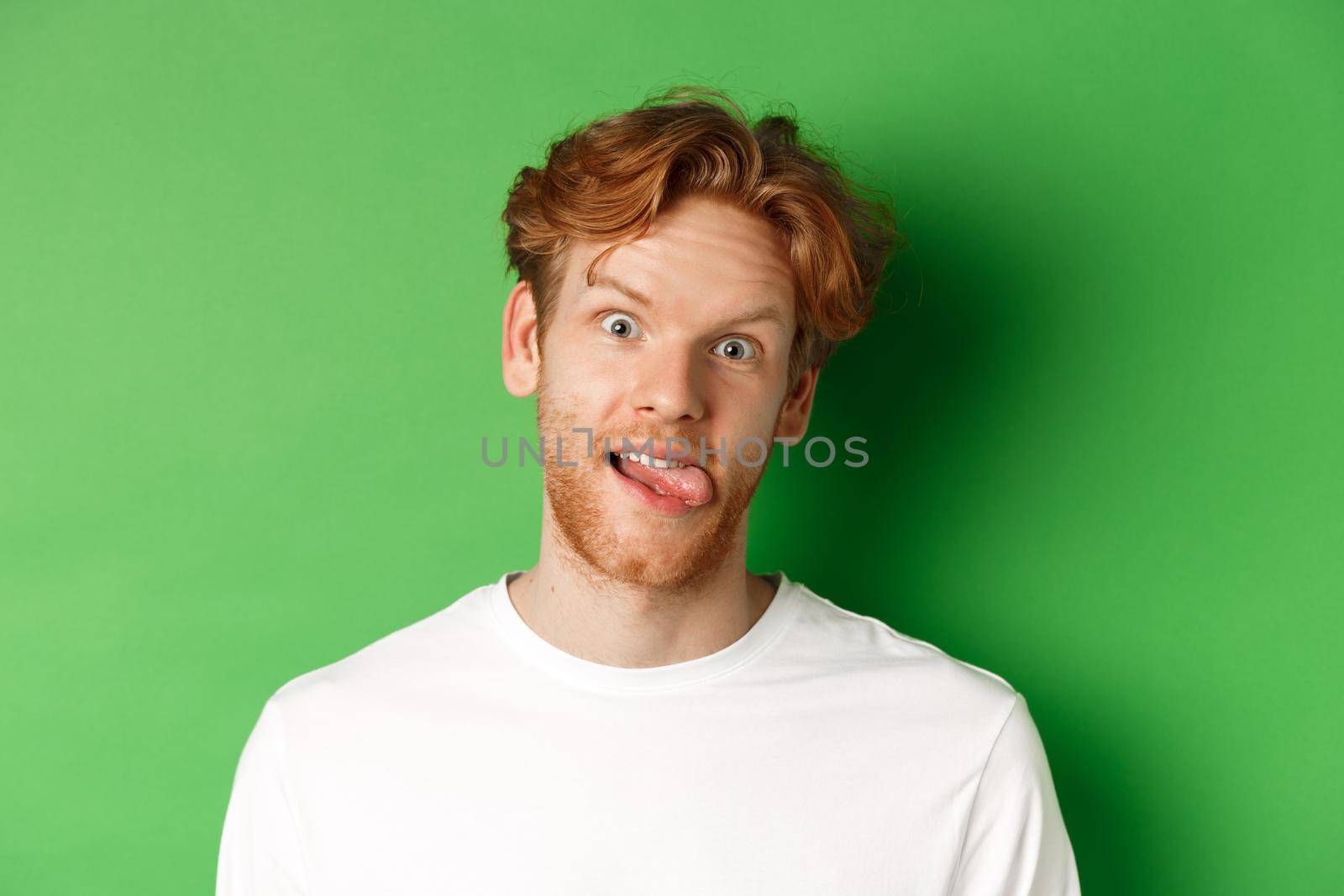 Emotions and fashion concept. Close up of funny redhead man showing silly faces, sticking tongue and staring at camera, standing over green background.