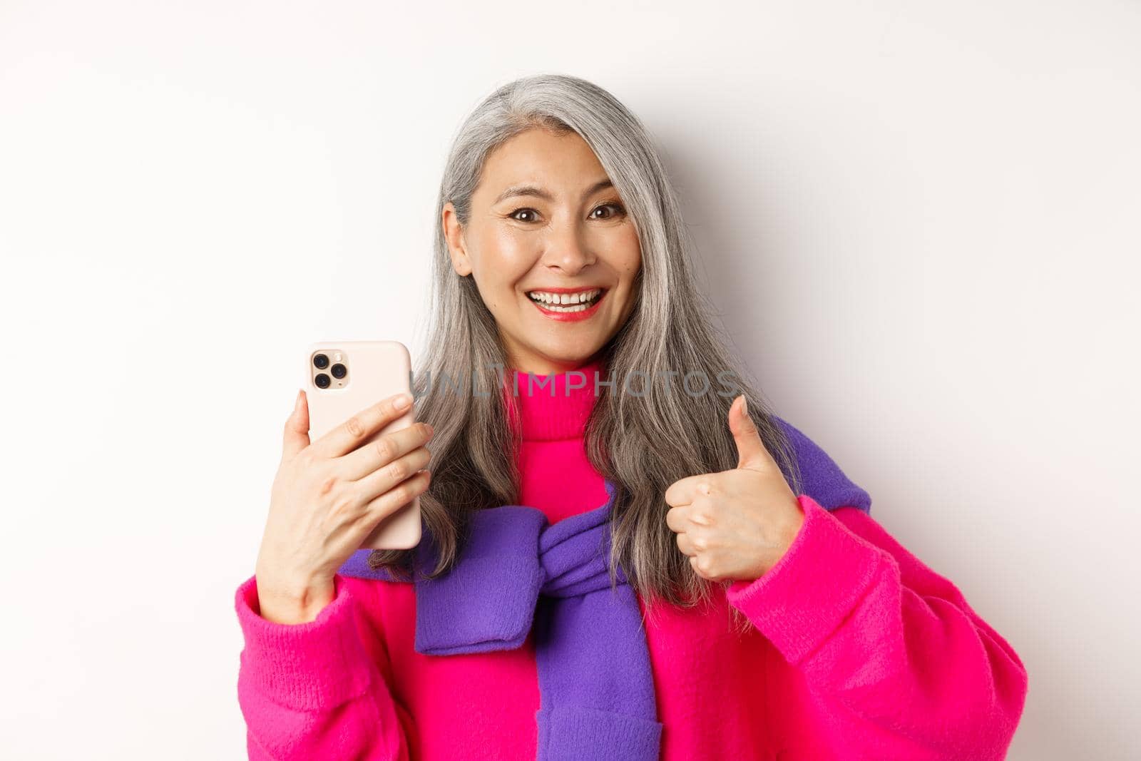 Online shopping. Close up of fashionable asian senior woman showing thumbs-up, using smartphone and approve something, standing over white background by Benzoix