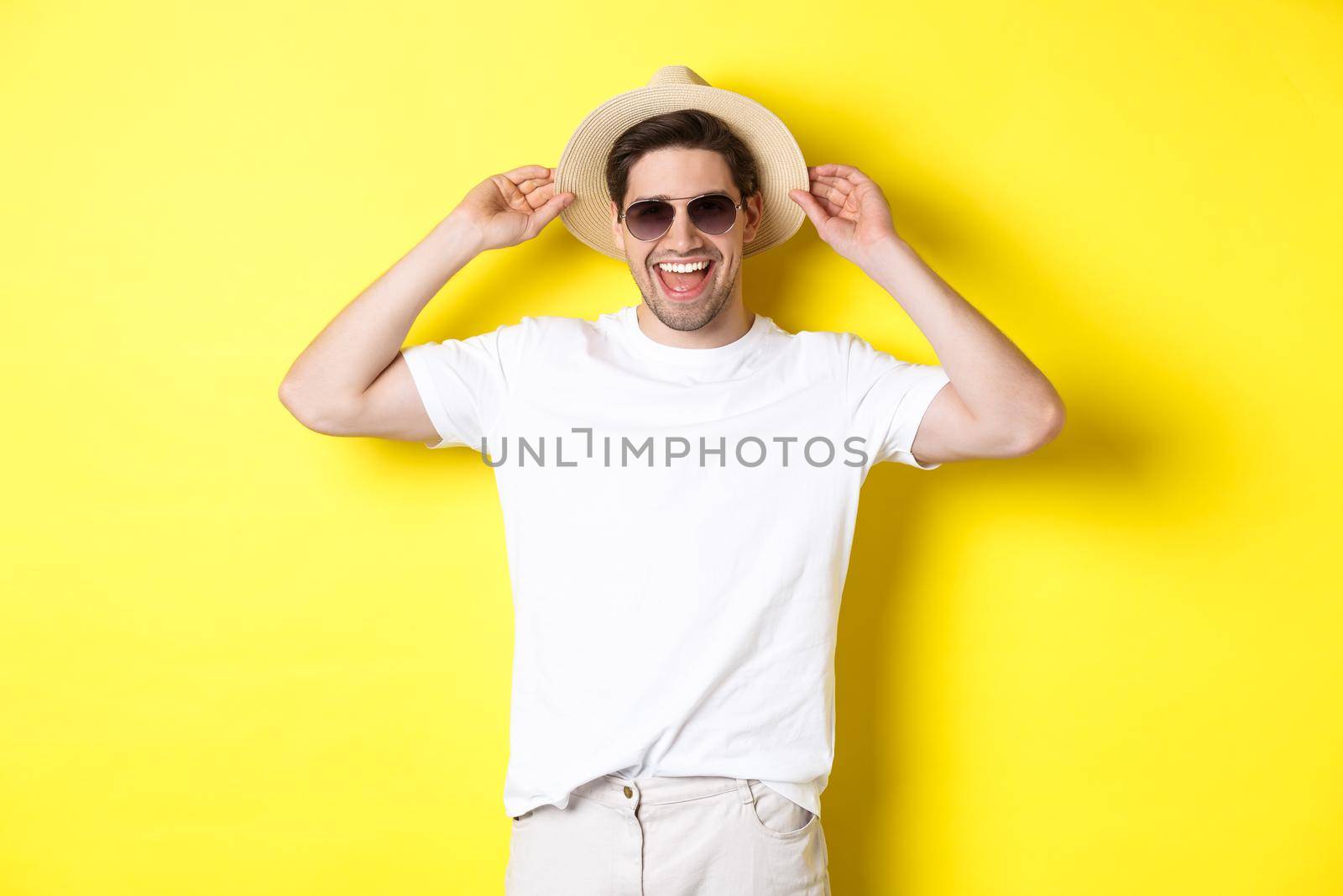Happy man on vacation, wearing straw hat and sunglasses, smiling while standing against yellow background.