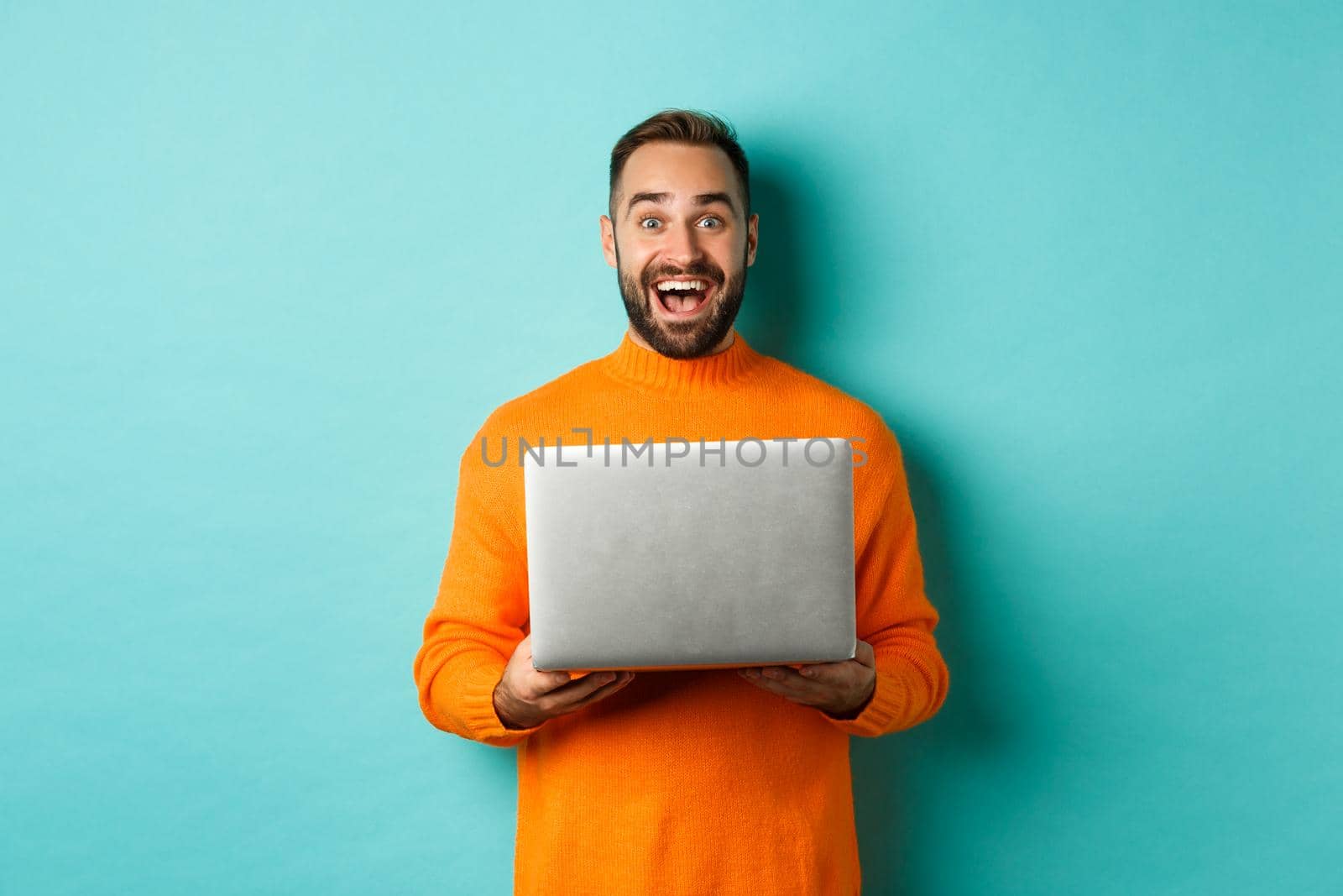 Happy man using laptop and looking excited at camera, standing with computer against light blue background.