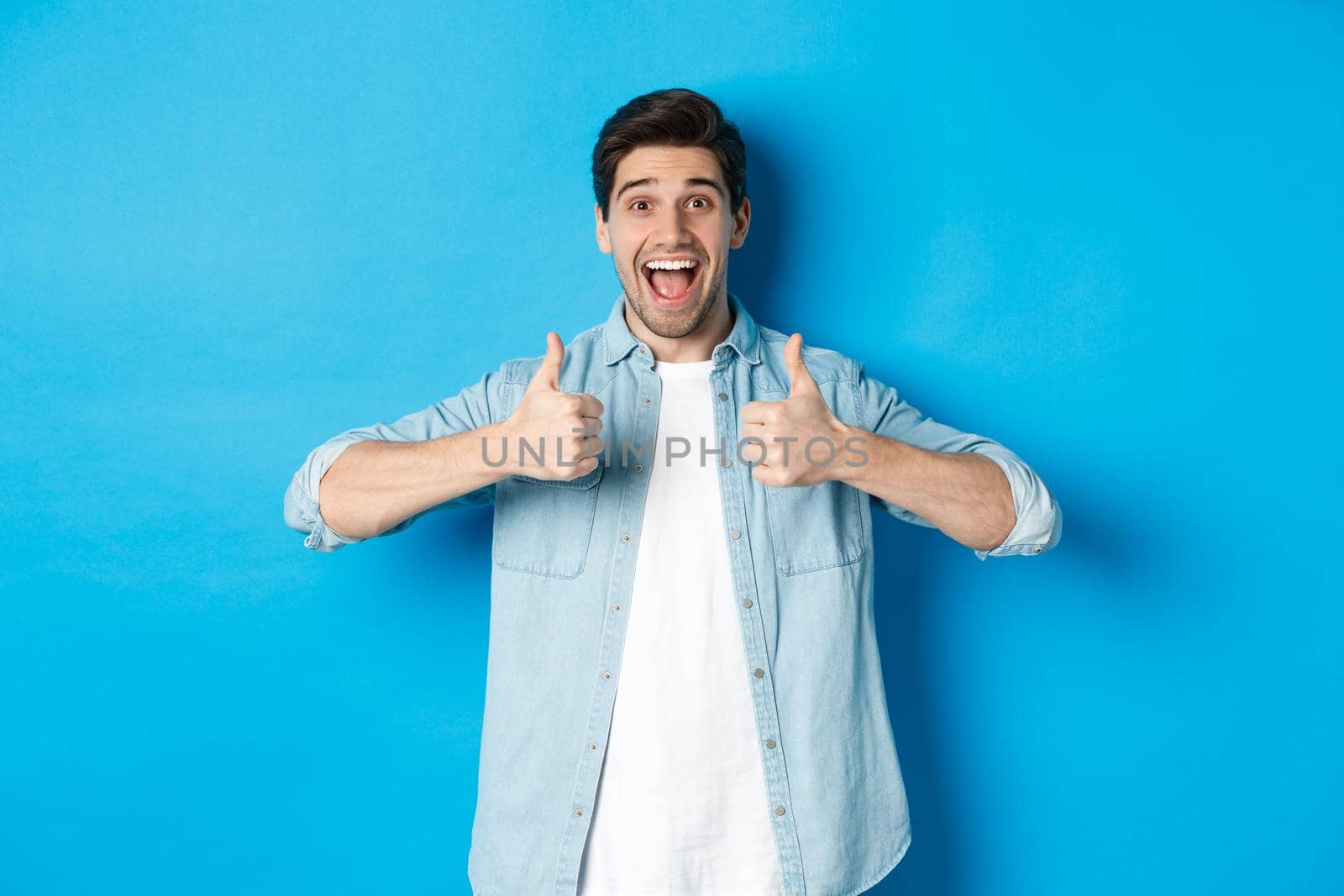 Smiling adult man showing thumbs up with excited face, like something awesome, approving product, standing against blue background.