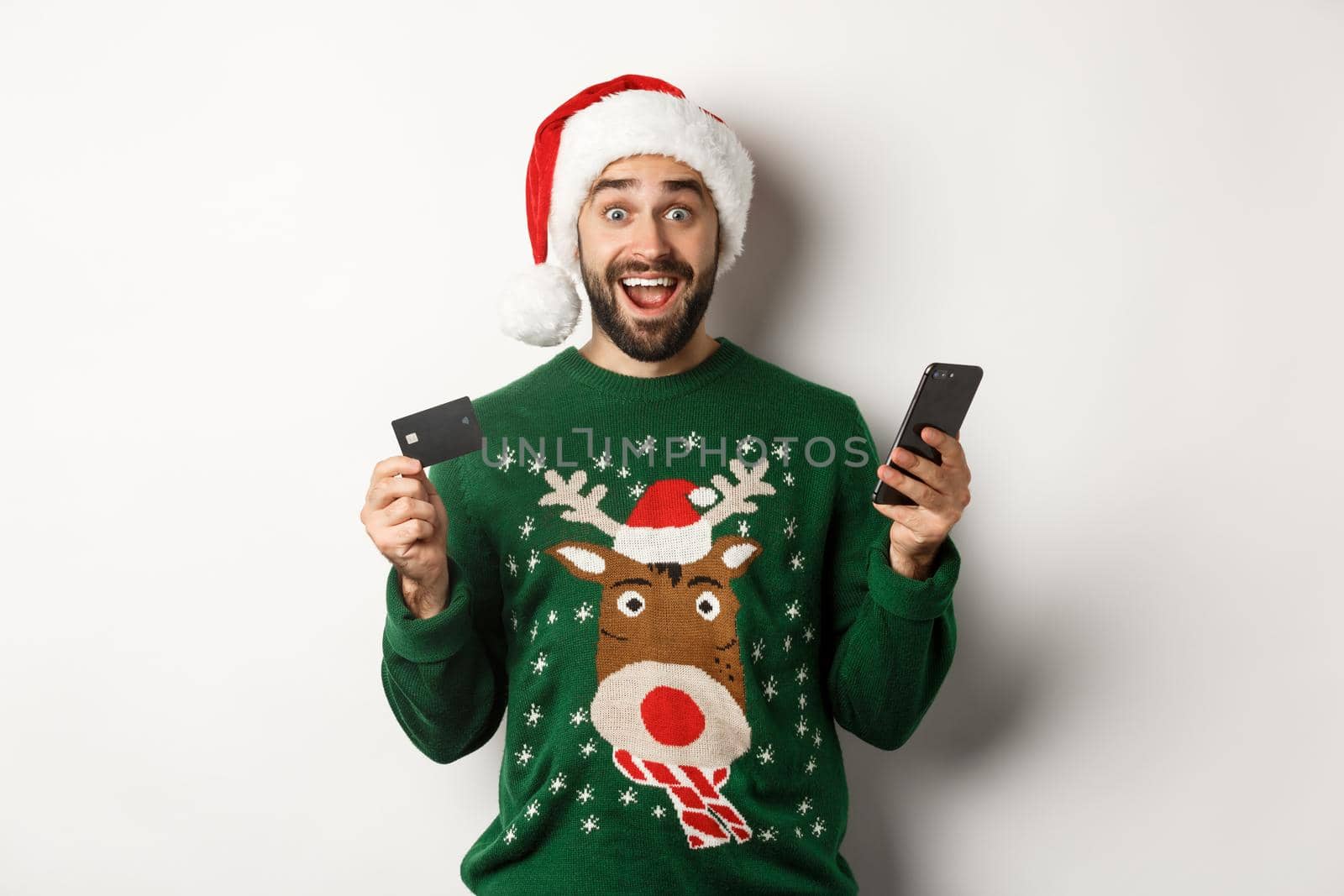 Online shopping and winter holidays concept. Surprised man in Santa hat, holding mobile phone and credit card, standing in sweater over white background.