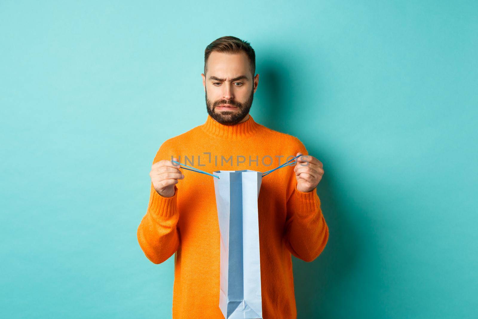 Disappointed man open shopping bag and dislike gift, frowning displeased, standing in orange sweater against turquoise background.