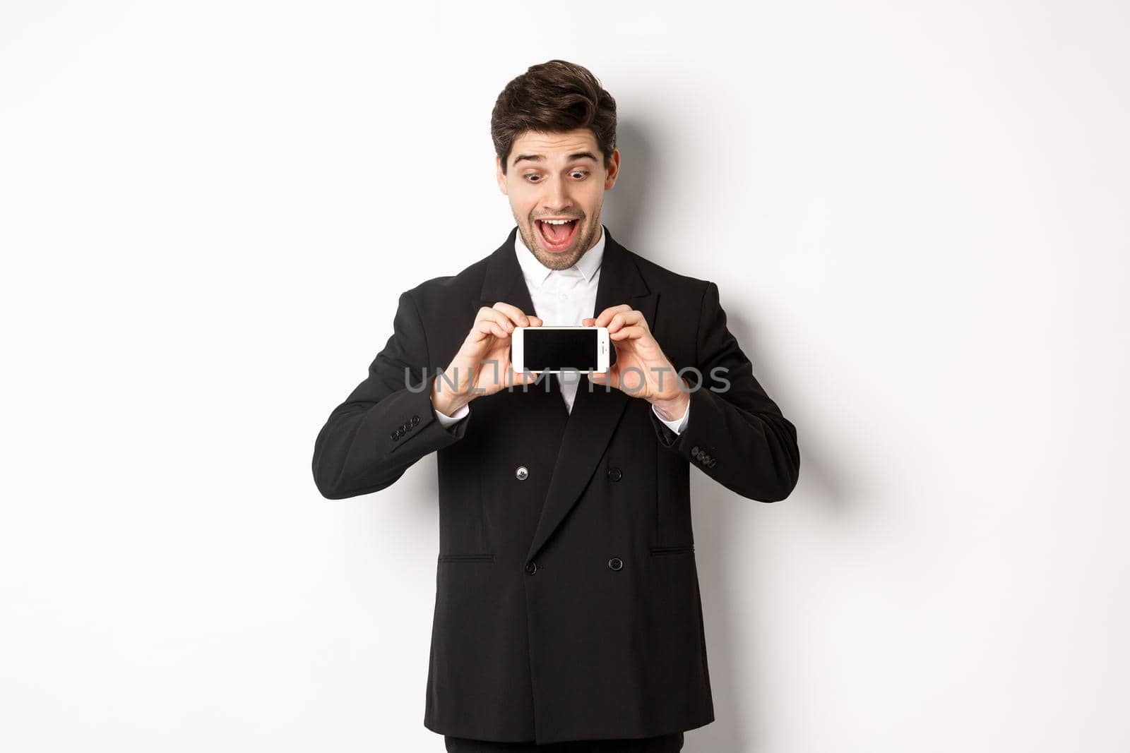 Portrait of excited handsome man showing smartphone screen, looking amazed at display with advertisement, standing over white background in black suit by Benzoix