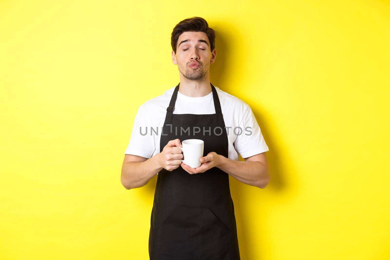 Man in black apron bringing cup of coffee and waiting for kiss, standing over yellow background by Benzoix