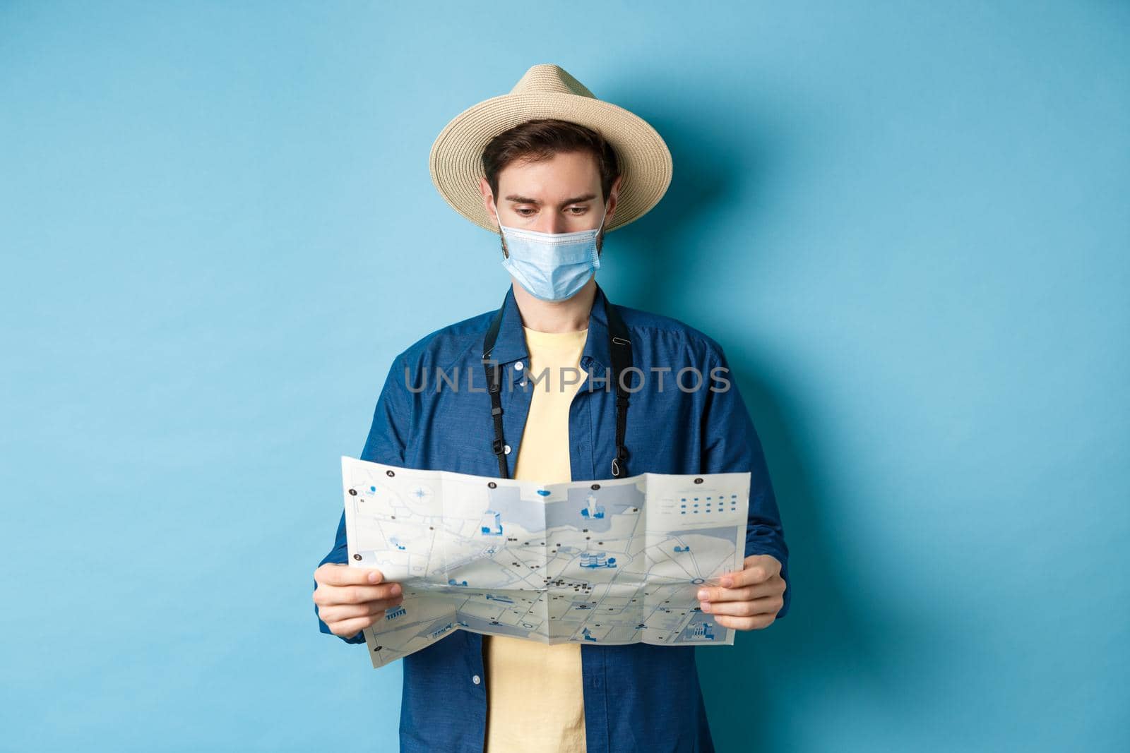 Covid-19, pandemic and travel concept. Young man in medical mask reading tourist map, searching for route to sightseeings, standing on blue background by Benzoix