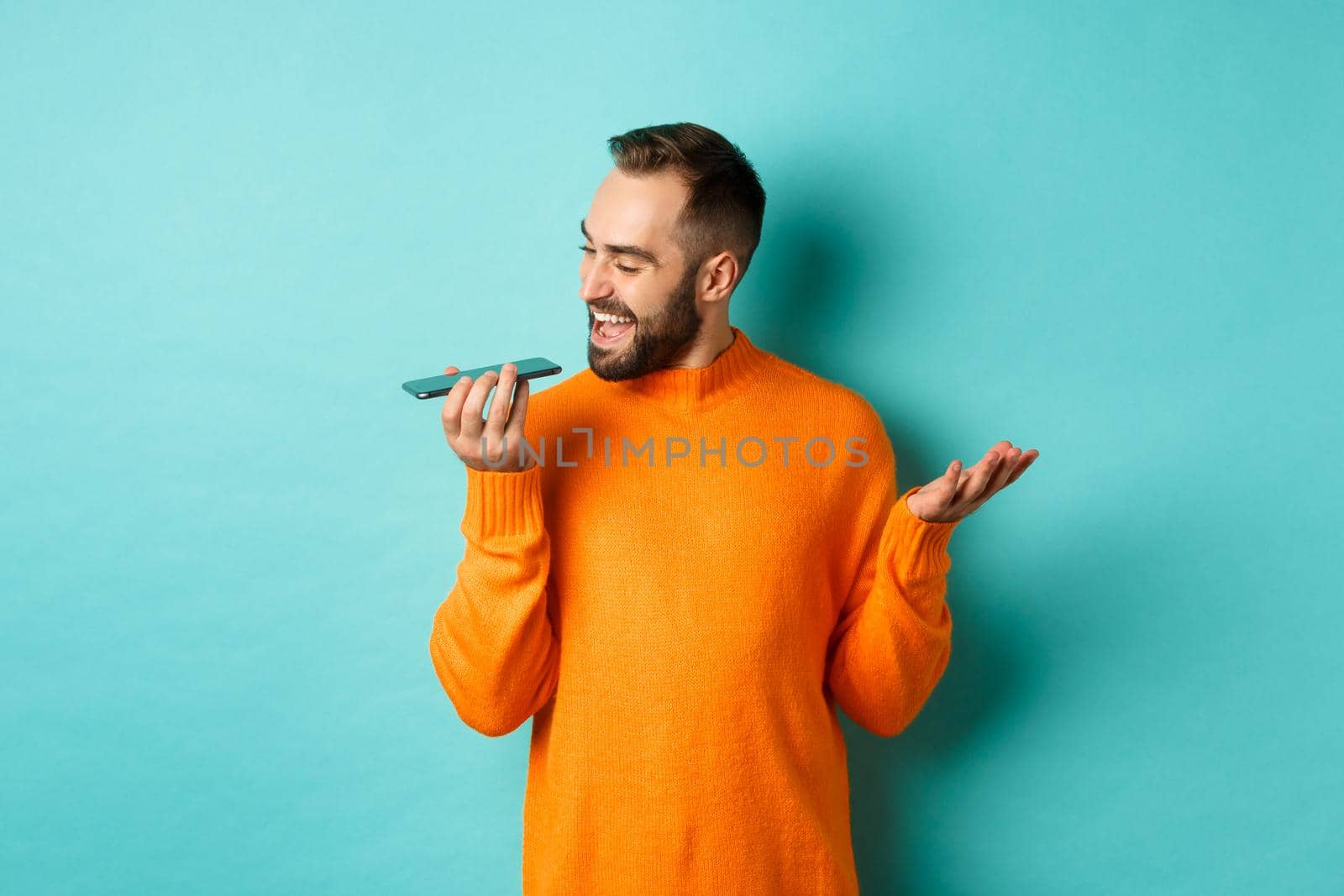 Happy man talking on speakerphone, gesturing and recording voice message on mobile phone, standing in orange sweater over light blue background.