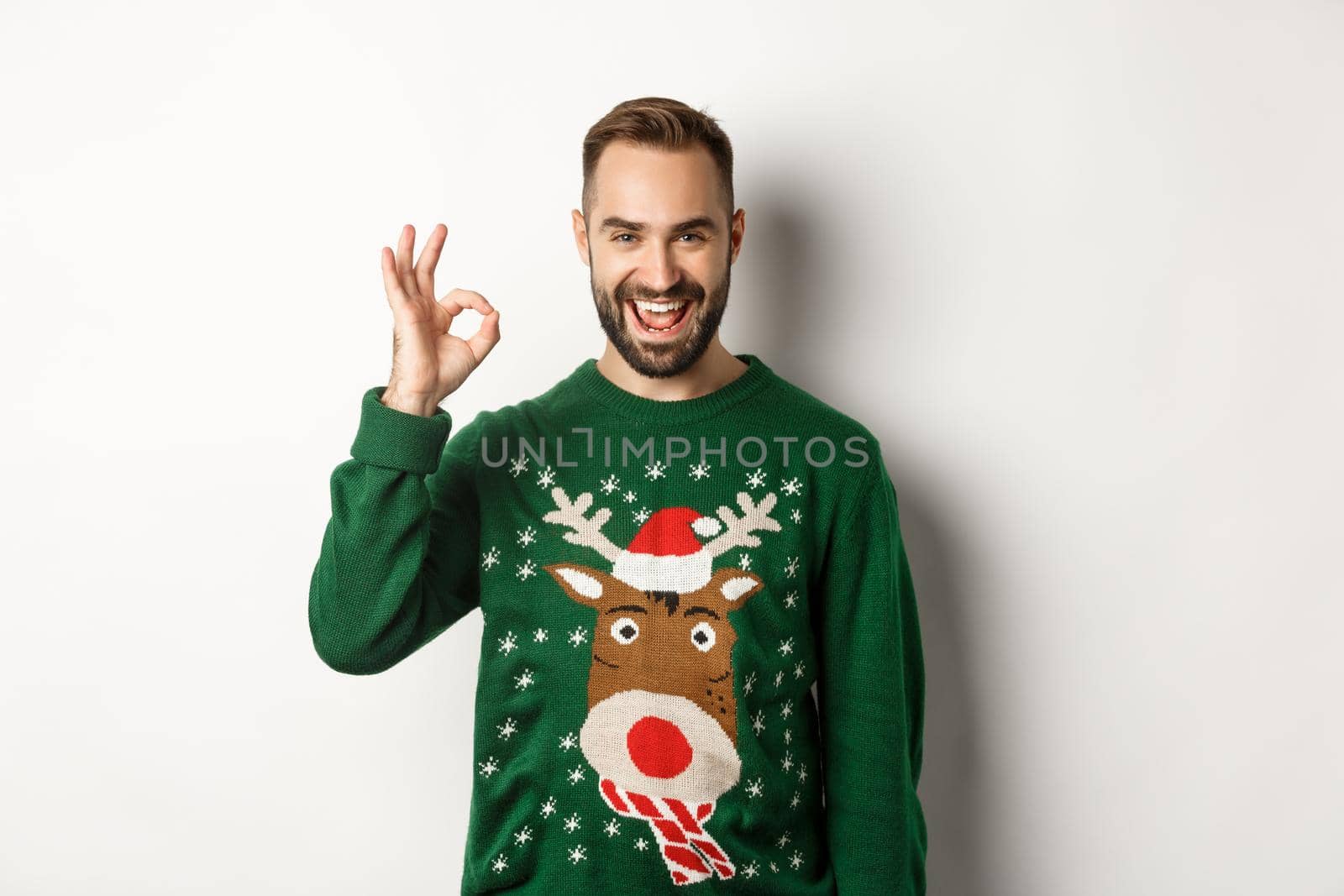 Winter holidays and christmas. Satisfied bearded man in green sweater, showing OK sign in approval, like something good, standing over white background.