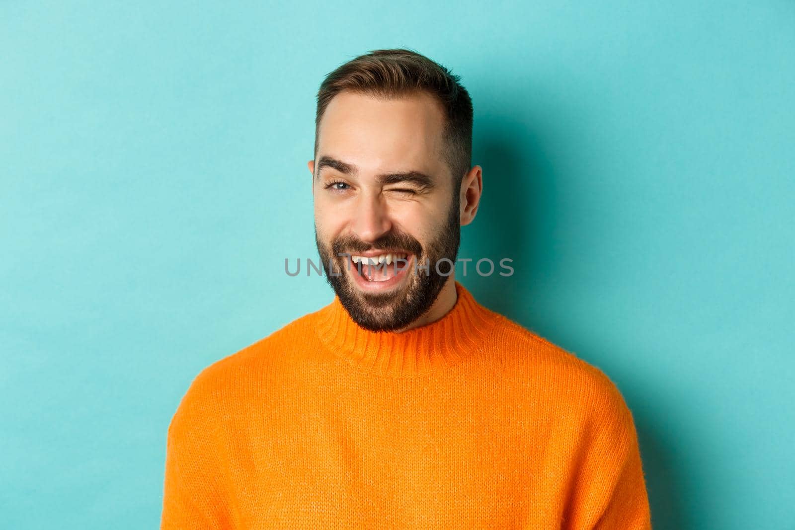 Handsome confident man winking at camera, smiling sassy, standing in orange sweater against blue background.