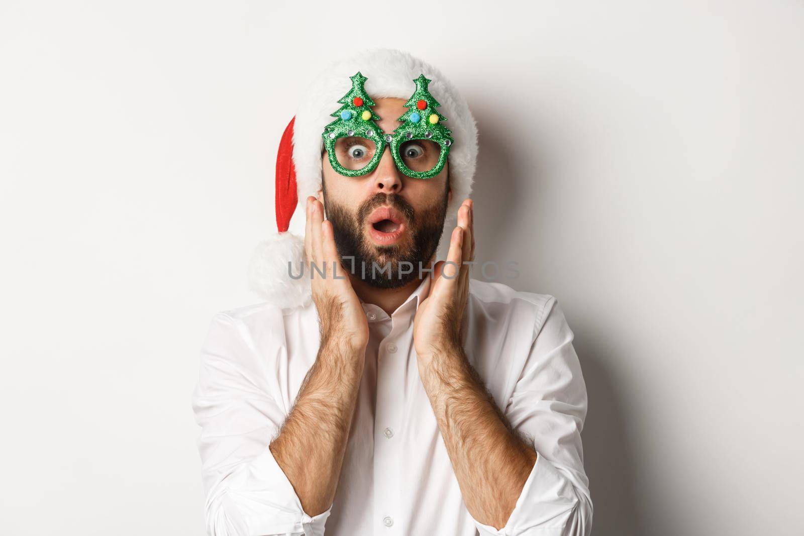 Adult man celebrating winter holidays, wearing christmas party glasses and santa hat, looking surprised at camera, standing over white background.