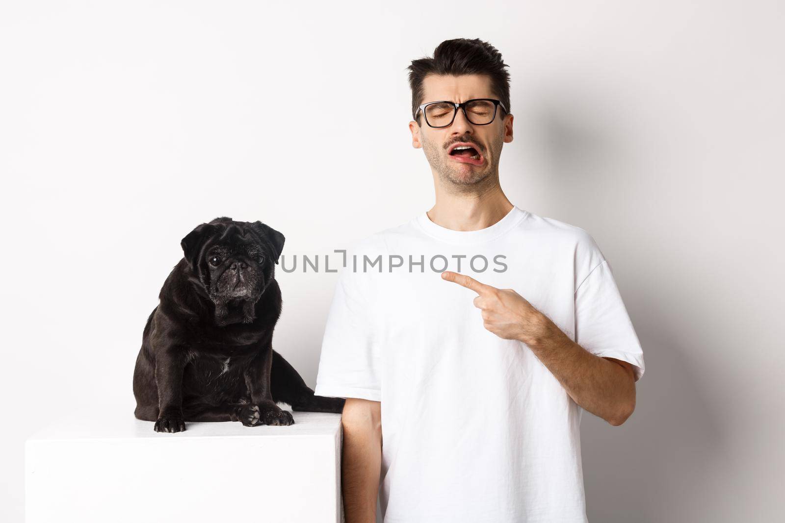 Upset crying man pointing at cute black pug and sobbing, complaining on his pet, standing sad against white background by Benzoix