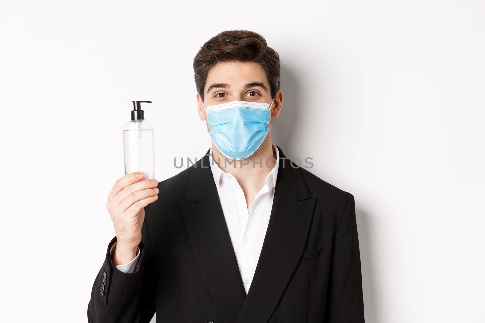 Concept of covid-19, business and social distancing. Close-up of handsome man in trendy suit and medical mask, showing hand sanitizer, standing against white background by Benzoix