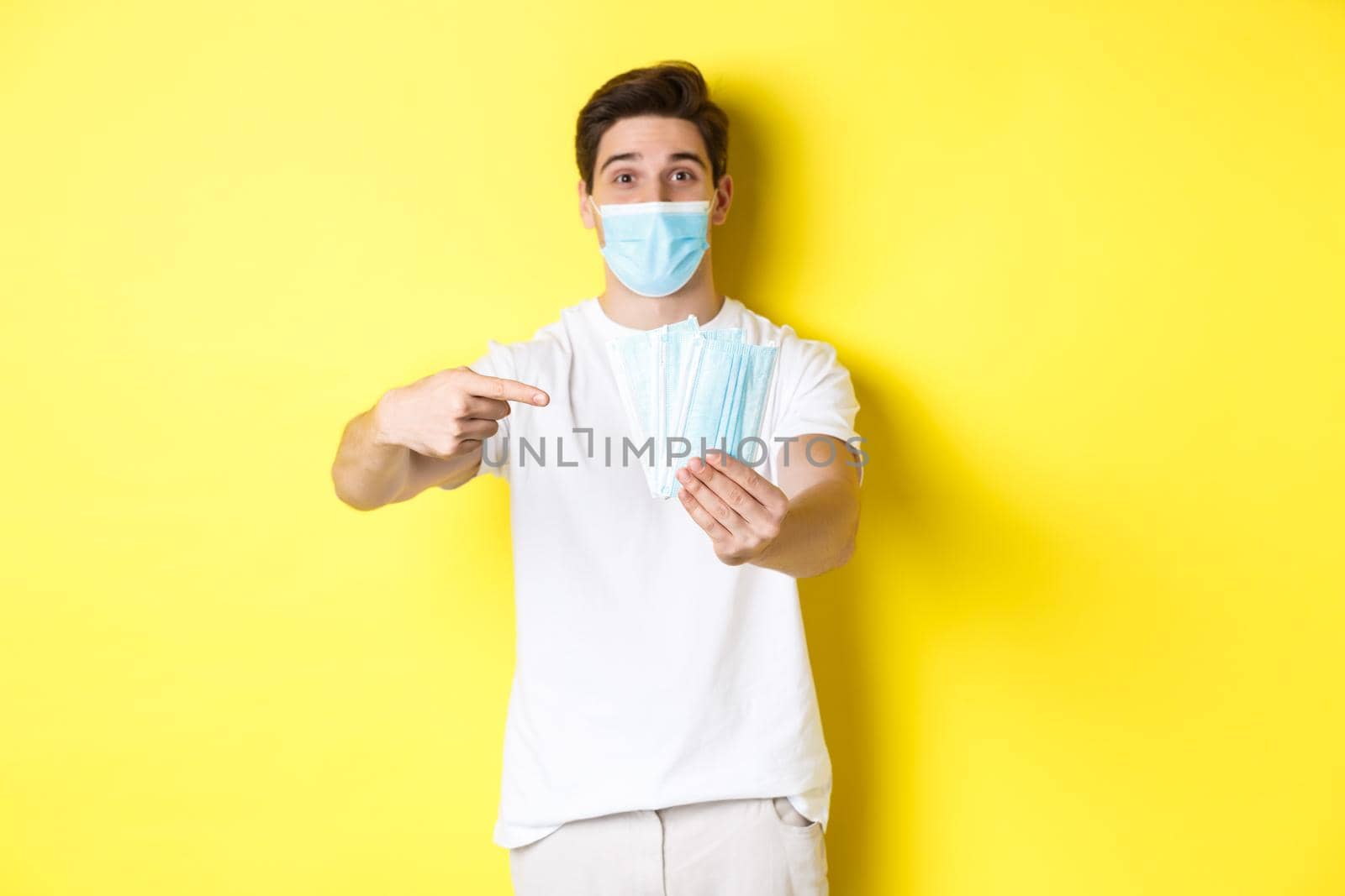 Concept of covid-19, quarantine and preventive measures. Young caucasian man giving medical masks for you, standing against yellow background.