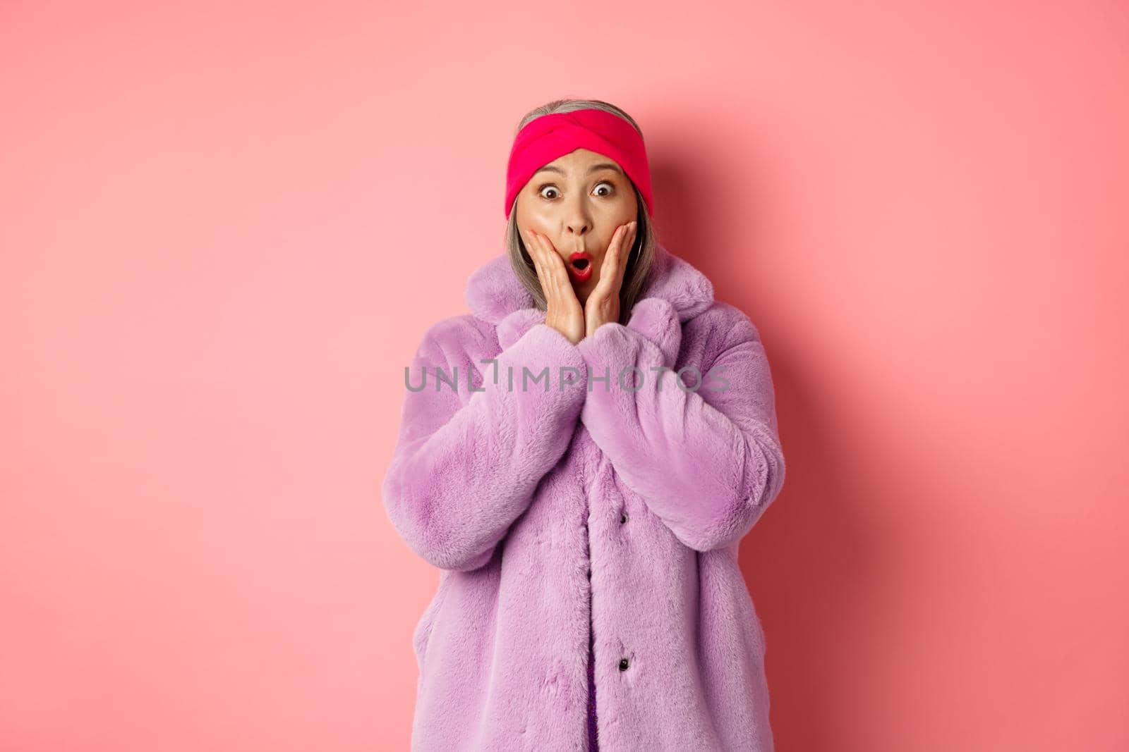 Fashion concept. Trendy old asian woman looking surprised at camera. Mature female in purple coat and headband looking amazed at camera, checking out promotion on pink background by Benzoix
