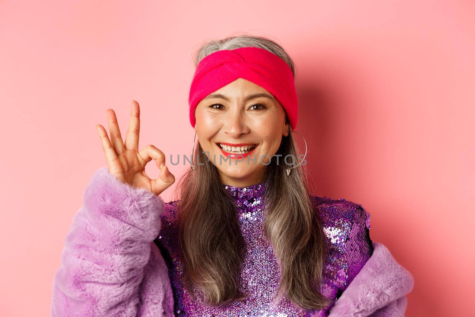 Fashion. Close-up of asian old lady in stylish disco outfit showing OK sign, approve and like something, smiling pleased, pink background.