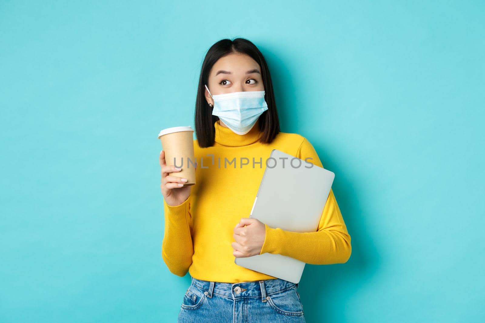 covid-19, health care and quarantine concept. Asian girl student in medical mask standing with laptop and coffee from cafe, standing over blue background.