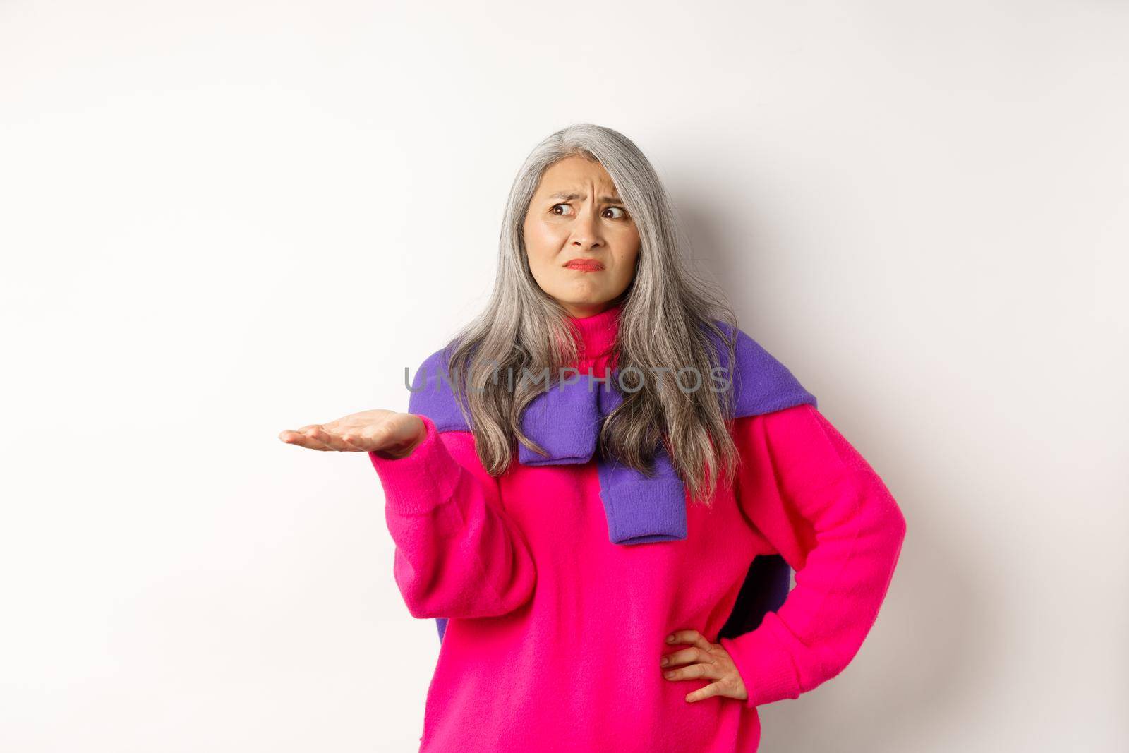 Funny asian mother with grey hair complaining, shrugging and looking left confused, pointing hand at something strange, standing over white background.