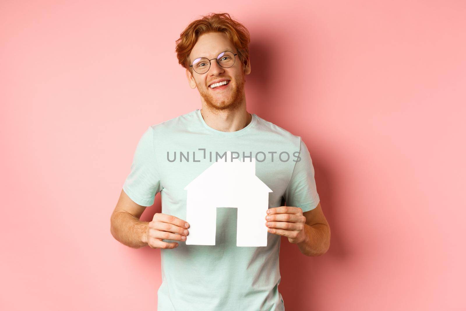 Real estate. Cheerful young man with red hair, wearing glasses and t-shirt, showing paper house cutout and smiling, buying apartment, pink background by Benzoix