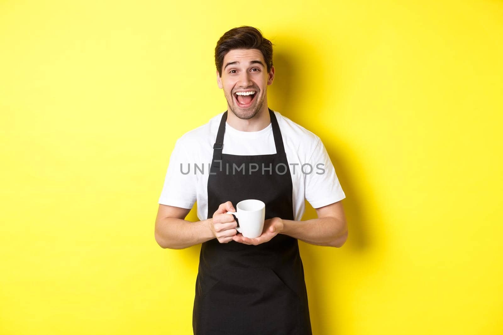 Happy barista in black apron holding coffee cup, laughing and standing over yellow background.