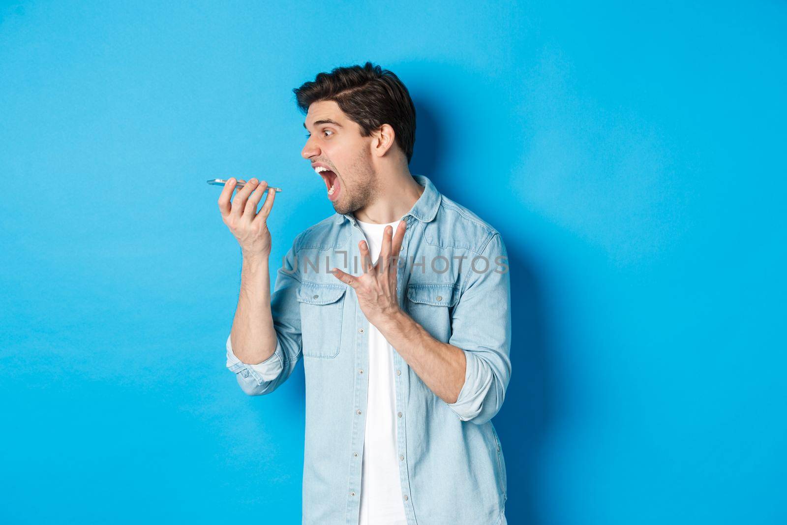 Angry and annoyed guy screaming at mobile phone, shouting on speakerphone while standing over blue background.