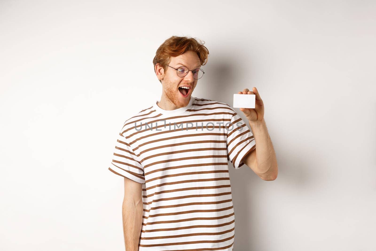 Shopping and finance concept. Excited young man with red hair, staring at plastic card and scream of joy, standing over white background.