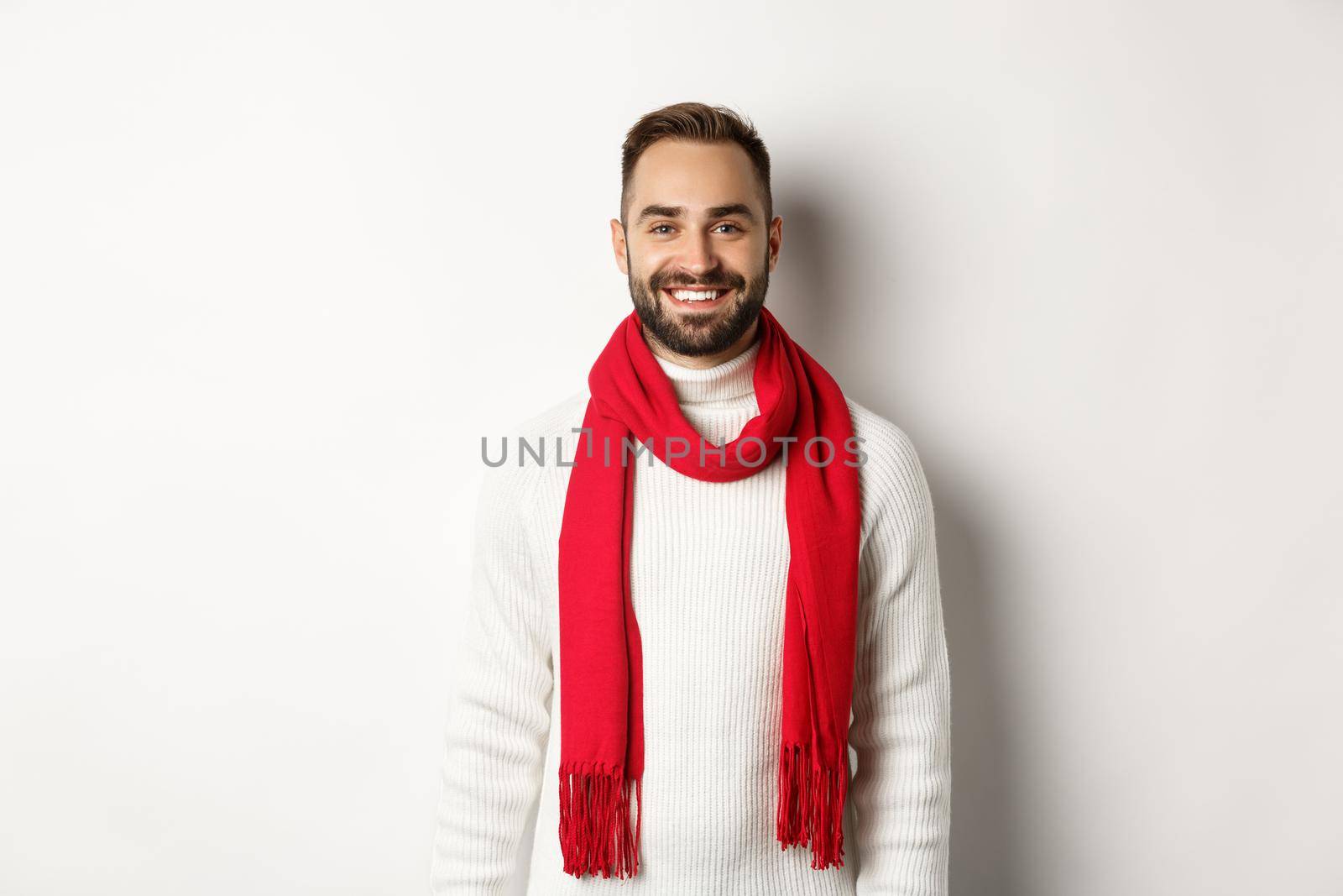 Winter holidays. Handsome adult man with red scarf looking happy at camera, standing in sweater against white background.