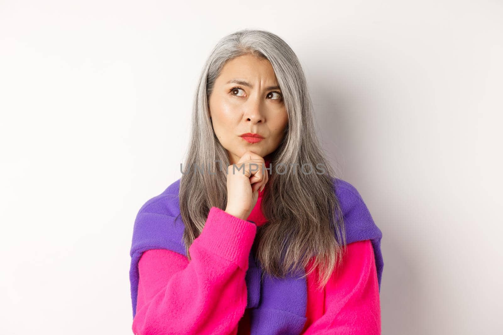 Close up of fashionable asian mature woman with grey hair, thinking and looking serious, looking aside while makign decision, standing over white background.