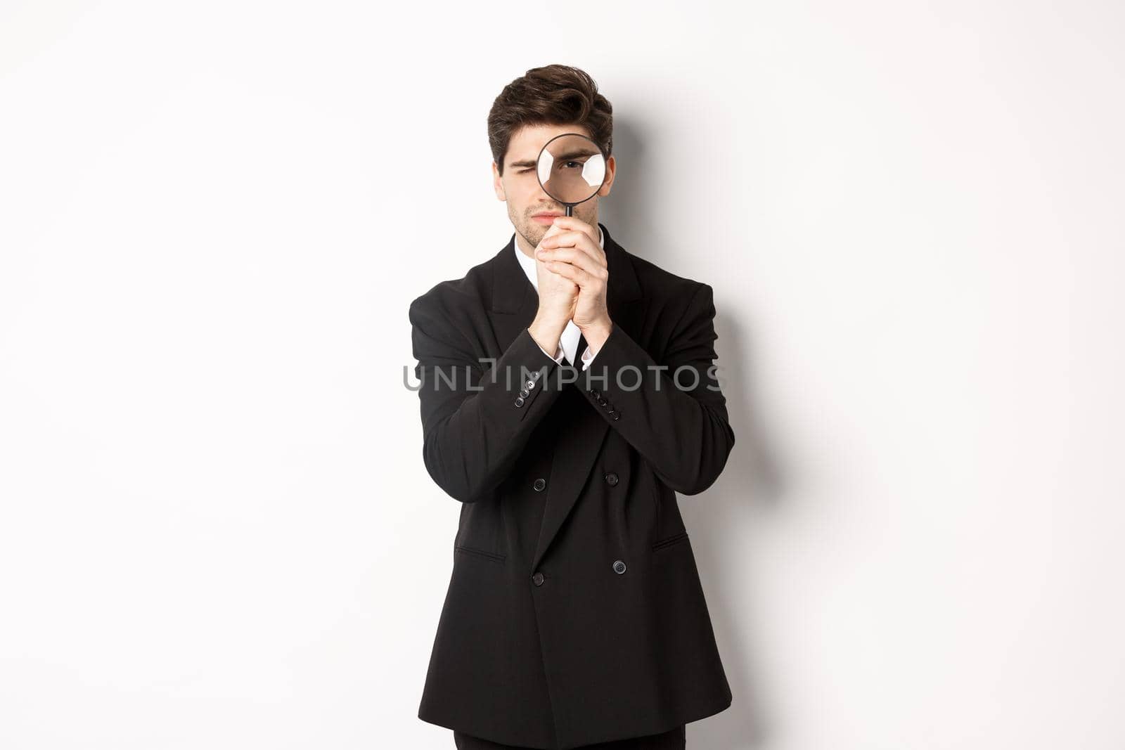 Image of serious businessman in black trendy suit, looking through magnifying glass, looking for employees, standing against white background by Benzoix