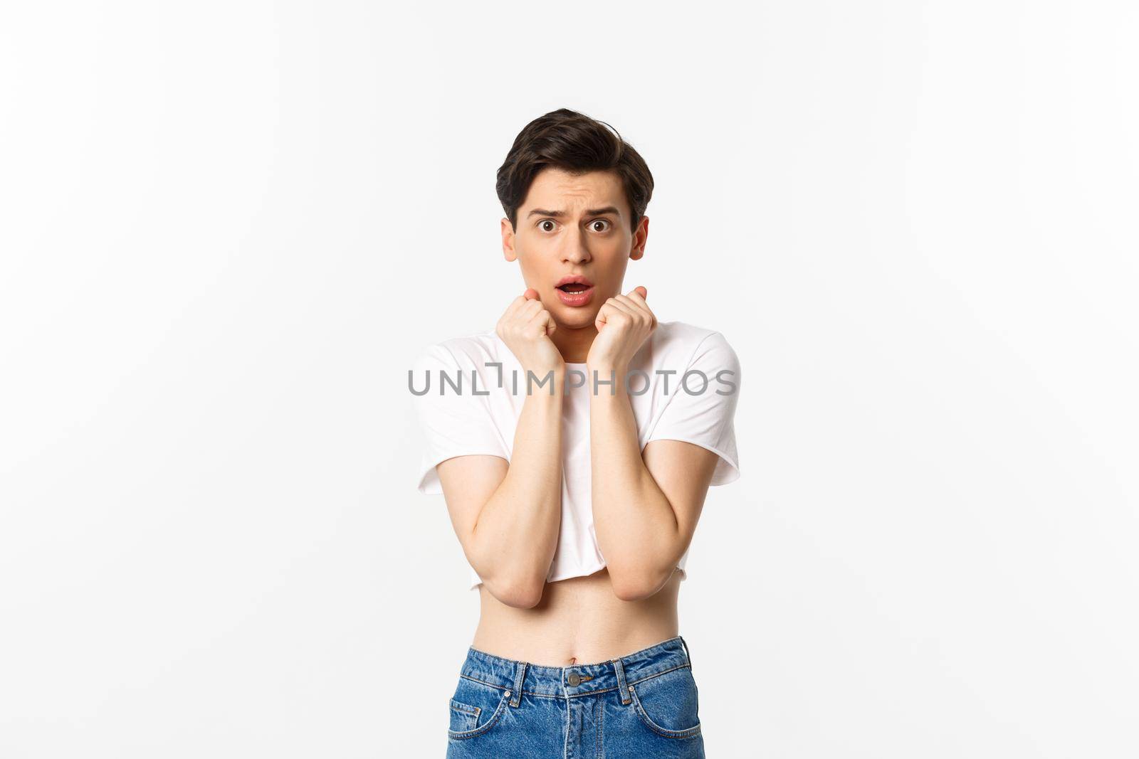 Image of scared androgynous man gasping and looking frightened, staring at something scary, standing in crop top against white background.