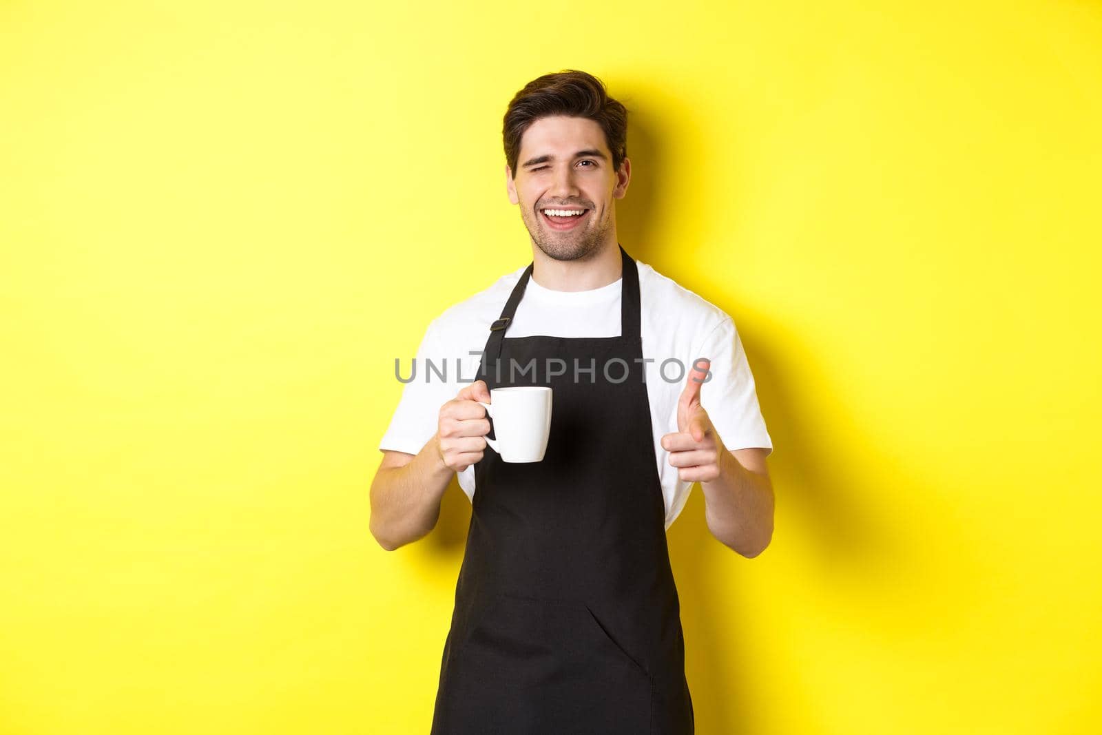 Handsome barista in black apron holding coffee cup, pointing finger at you, inviting visit cafe, standing over yellow background by Benzoix