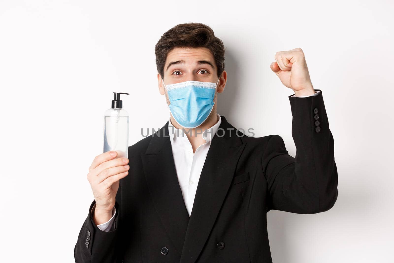 Concept of covid-19, business and social distancing. Close-up of happy man in trendy suit and medical mask, cheering and raising hand up, showing hand sanitizer, white background by Benzoix