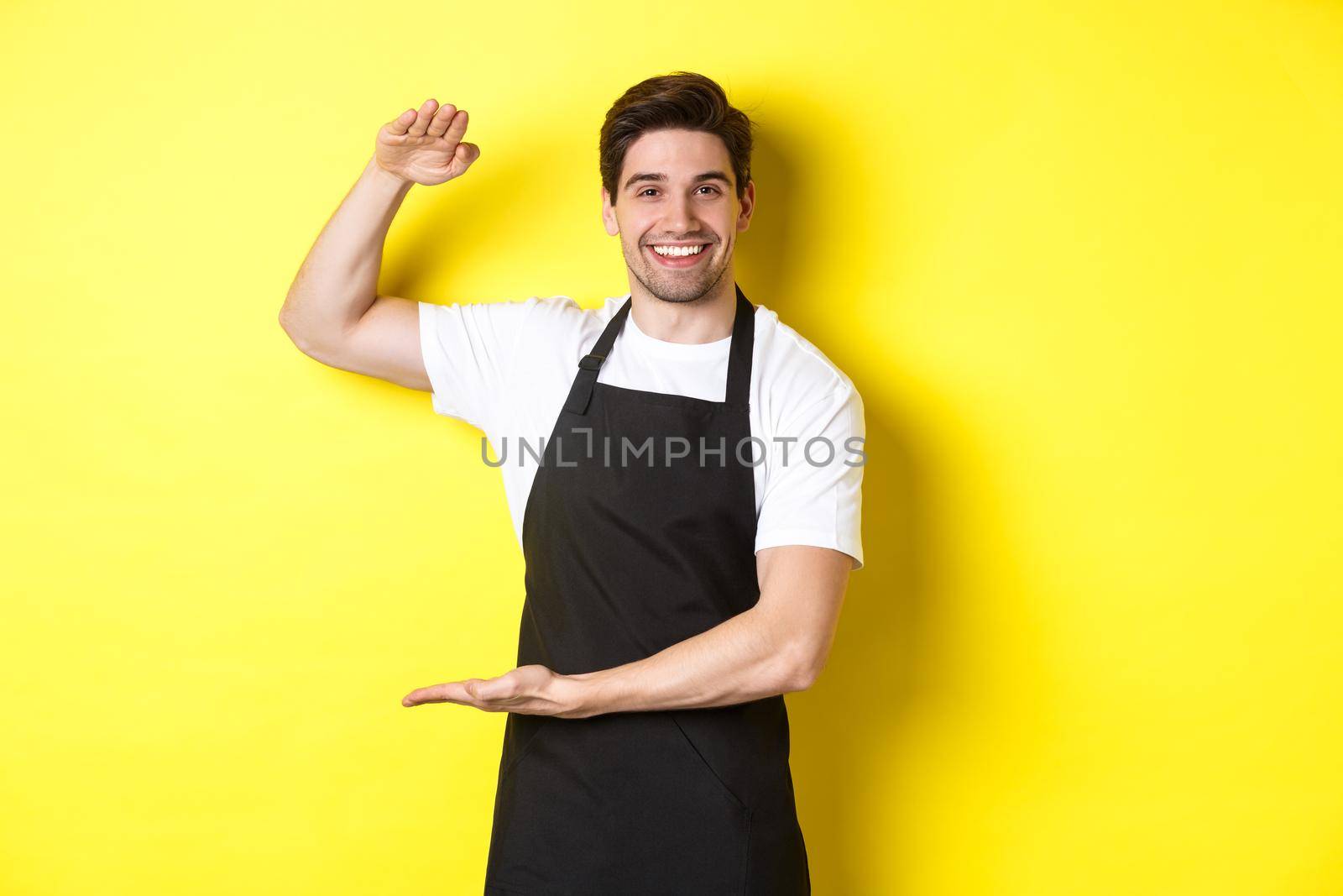 Smiling handsome barista showing something long or large, standing over yellow background by Benzoix