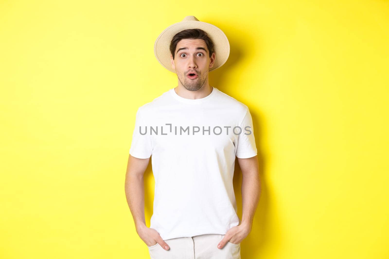 Concept of travelling and tourism. Surprised caucasian guy in straw hat, say wow, standing against yellow background by Benzoix