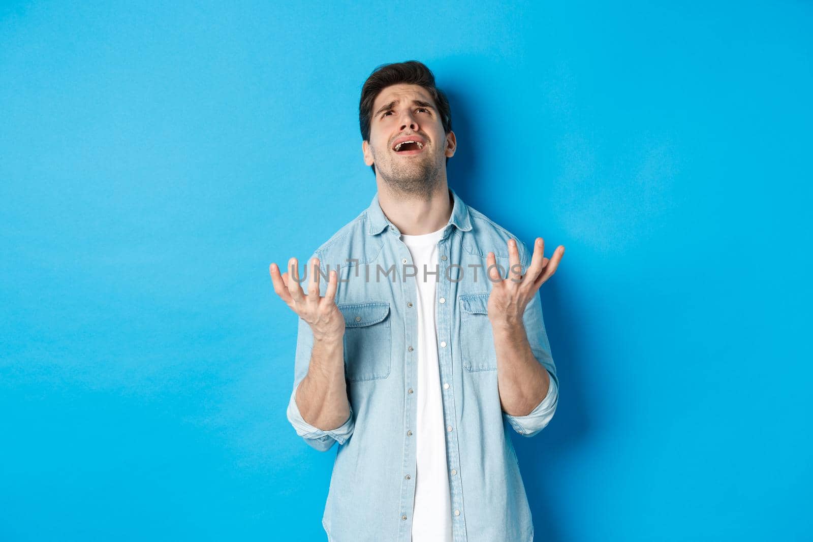 Frustrated guy complaining to god, looking up and pleading, standing against blue background upset.