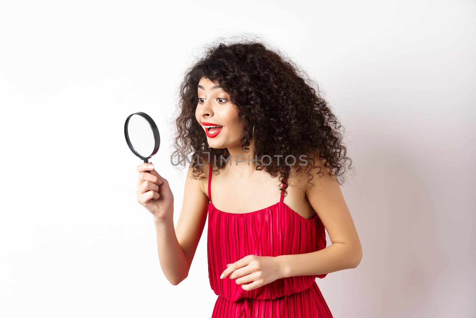 Beautiful woman in red dress and makeup looking at something with magnifying glass, checking out interesting promo, standing over white background.