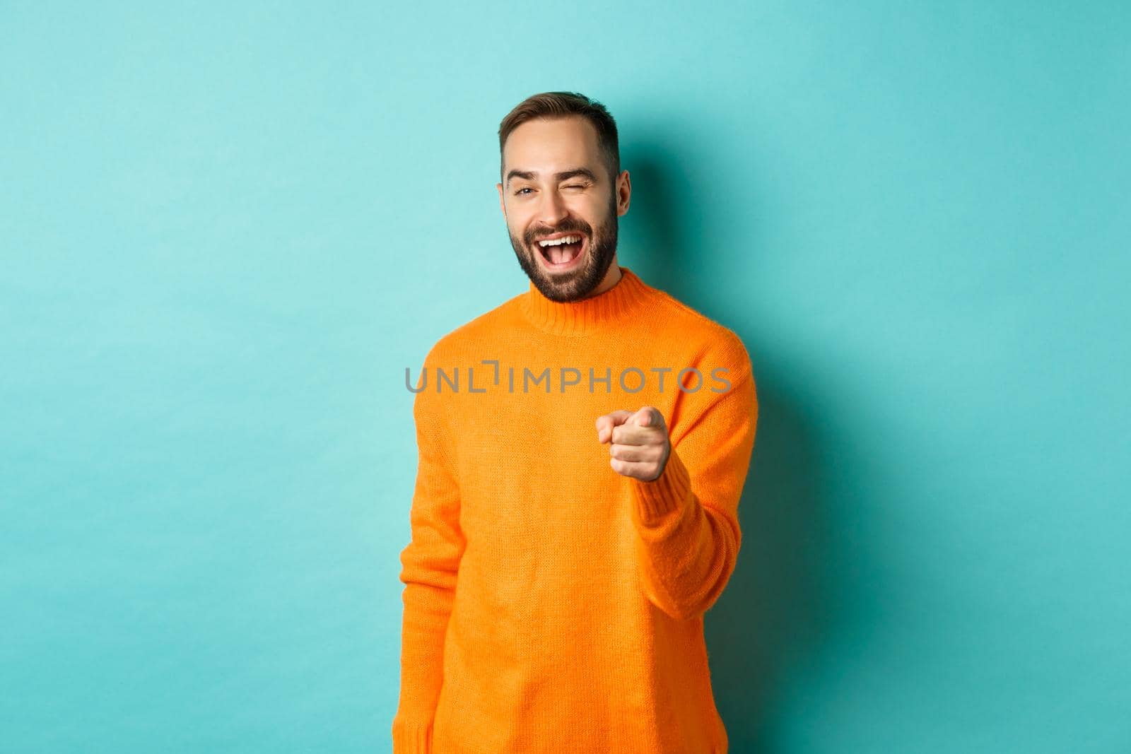 Cheeky attractive man congrats you, praising good job, winking and pointing at camera, standing over light blue background.