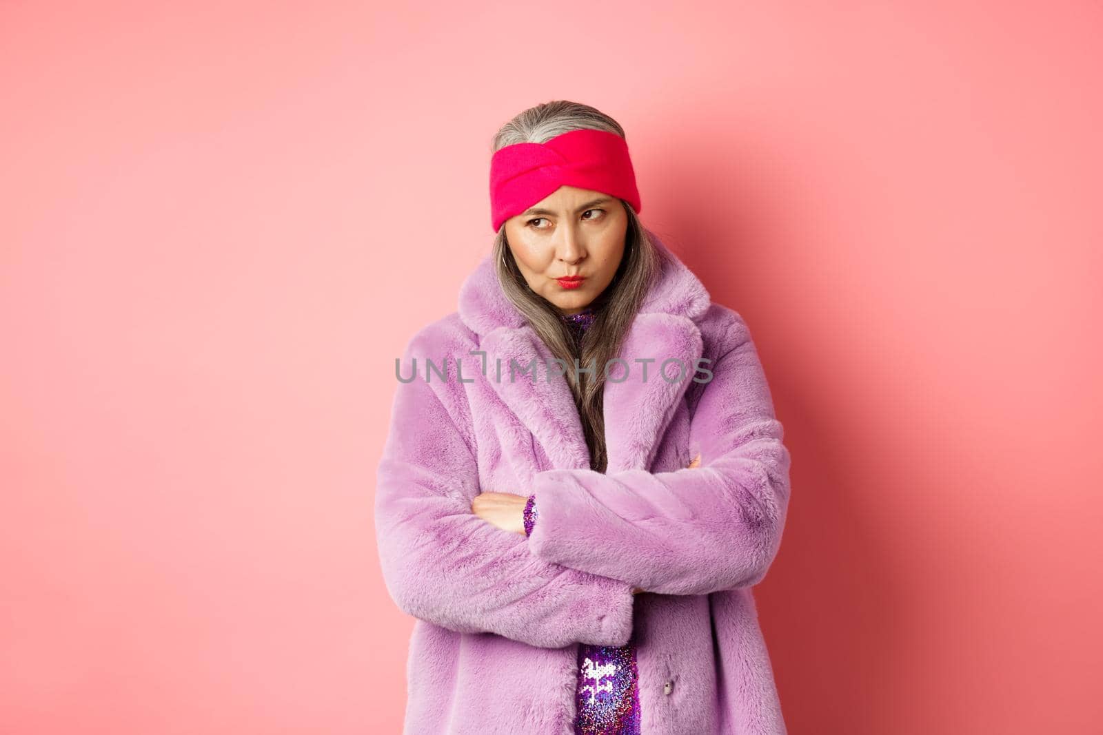 Grumpy funky old asian lady looking from under forehead, pouting and looking left with angry and disappointed face, standing in stylish purple faux fur coat against pink background.