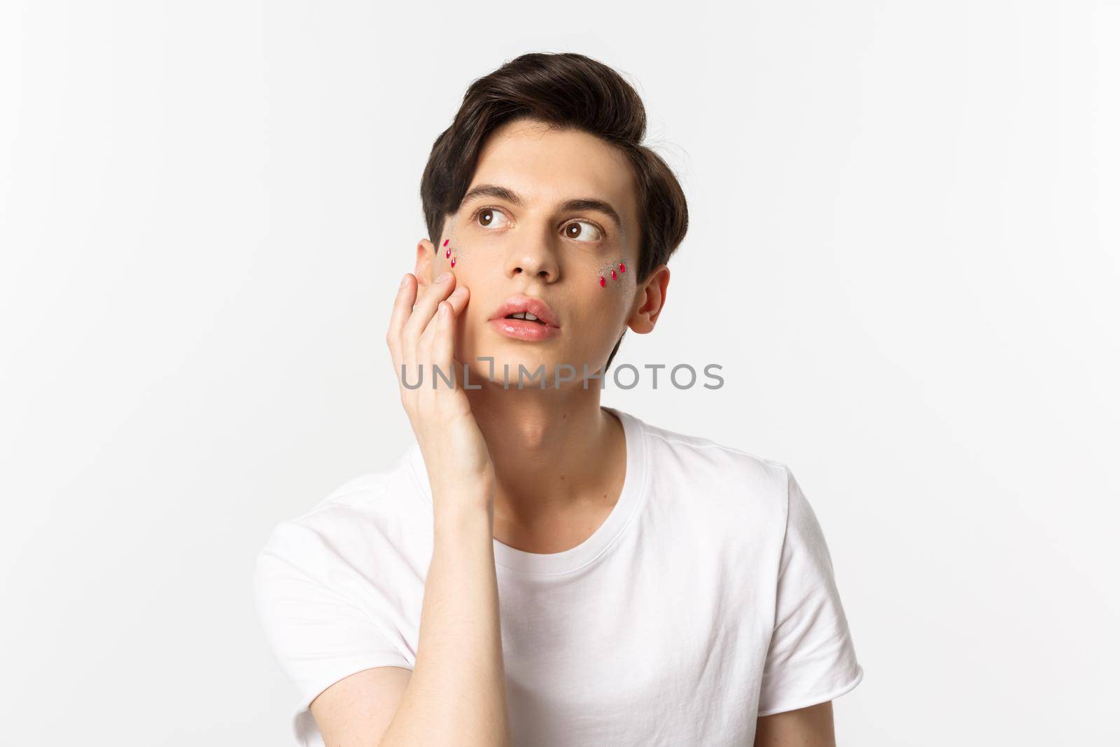 People, lgbtq community and lifestyle concept. Beautiful young gay man applying glitter under eyes for pride party, standing over white background.