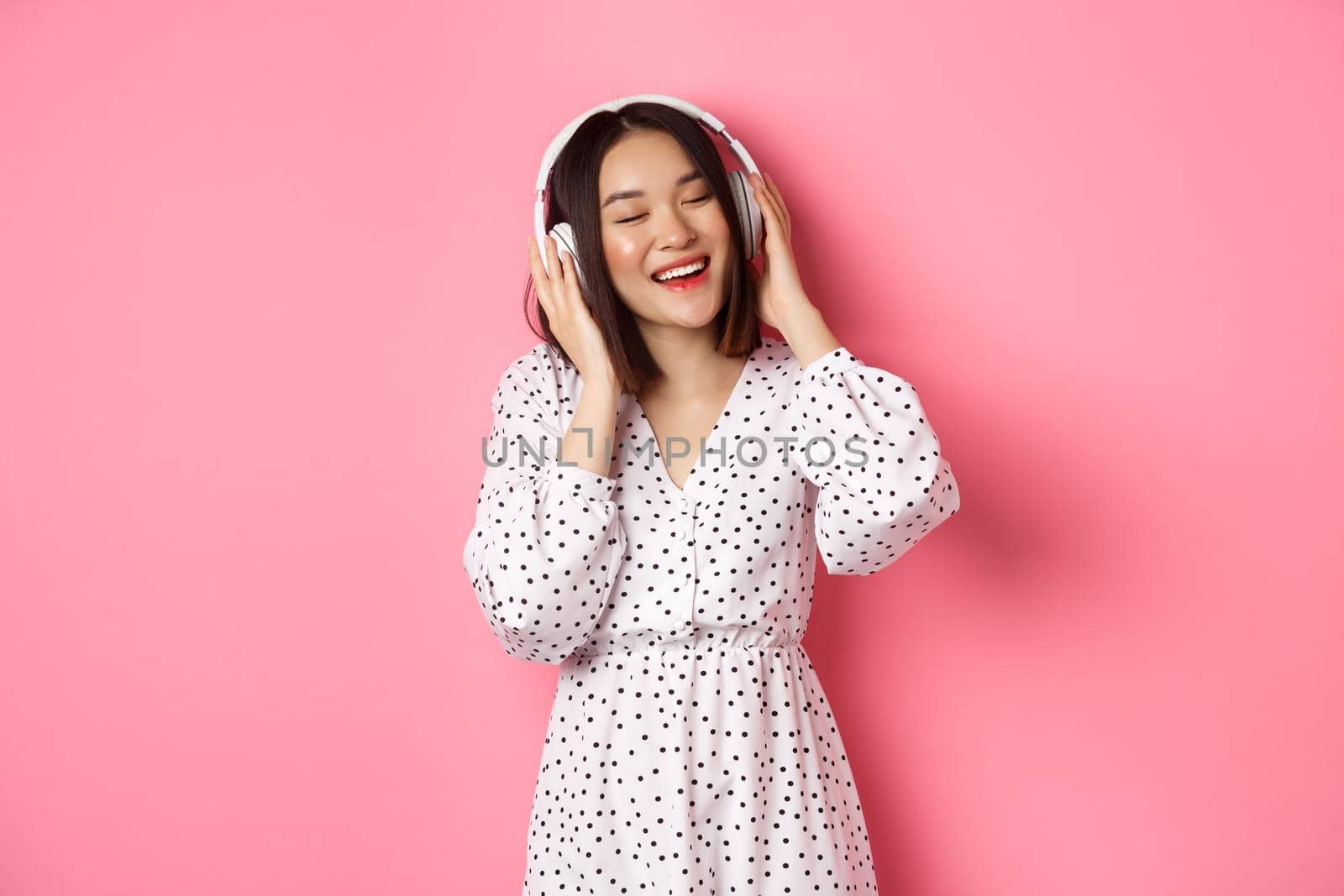Romantic asian woman smiling happy, listening music in headphones and dancing, standing in trendy dress over pink background.