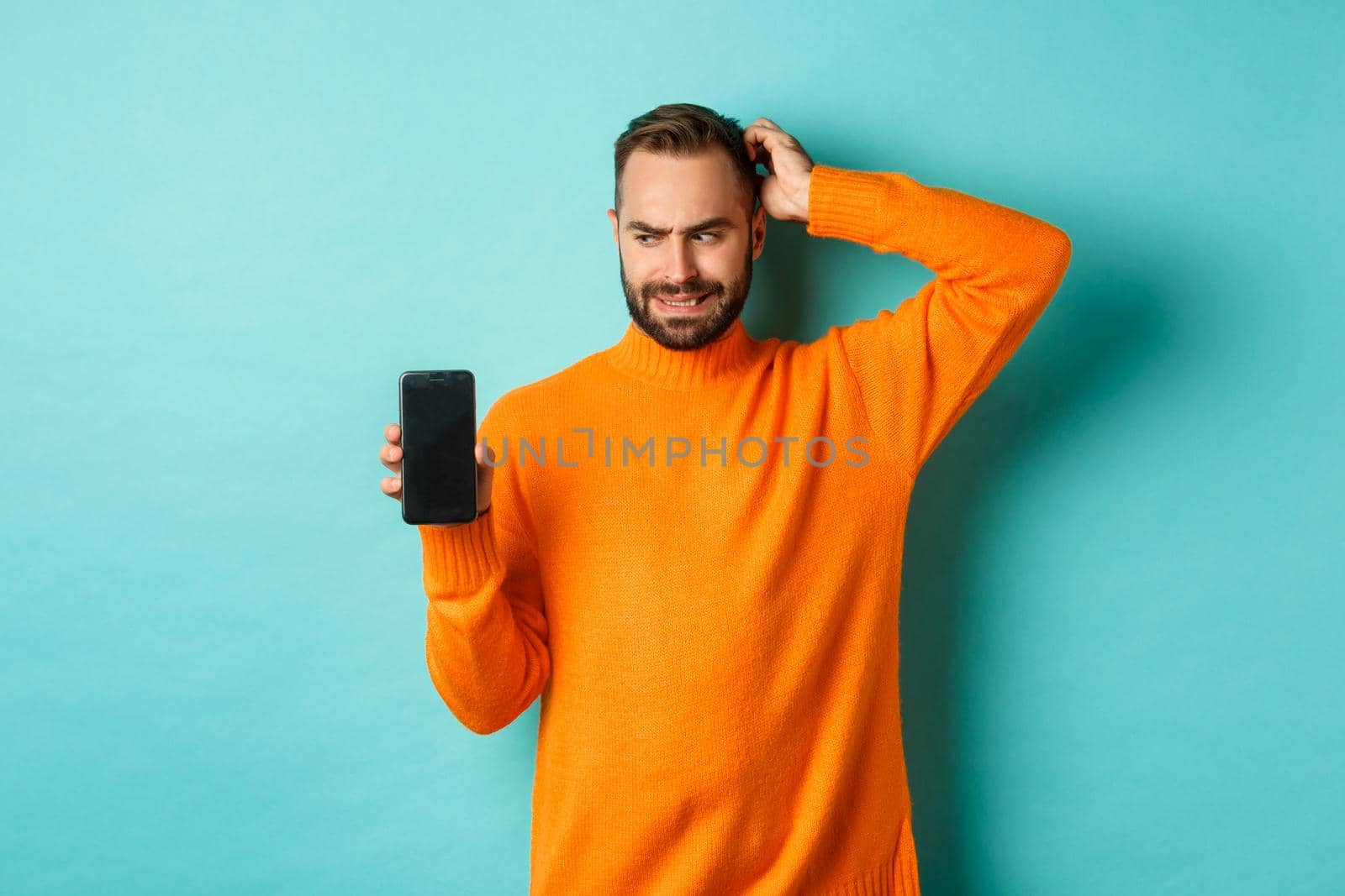 Confused guy scartching head and showing smartphone screen, looking puzzled, standing over turquoise background. Copy space