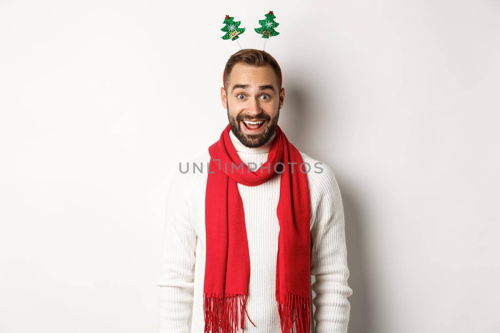 Christmas party and celebration concept. Happy bearded man looking surprised, wearing funny accessory hat, standing against white background.